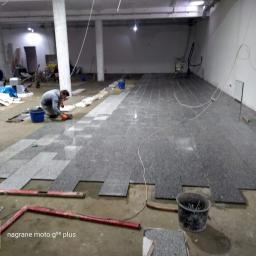 Daniel - Układanie szarej, granitowej podłogi w dużym, surowym pomieszczeniu; widoczny pracownik w pomarańczowych rękawicach, poziomica i wiadro z narzędziami.