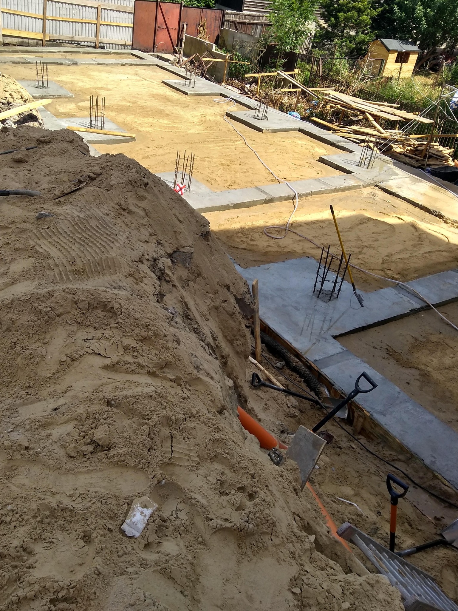 Wykop fundamentów z betonowymi ławami i wystającymi zbrojeniami, widoczny stos ziemi, szpadle i drabina, przygotowanie do wylewania fundamentów.