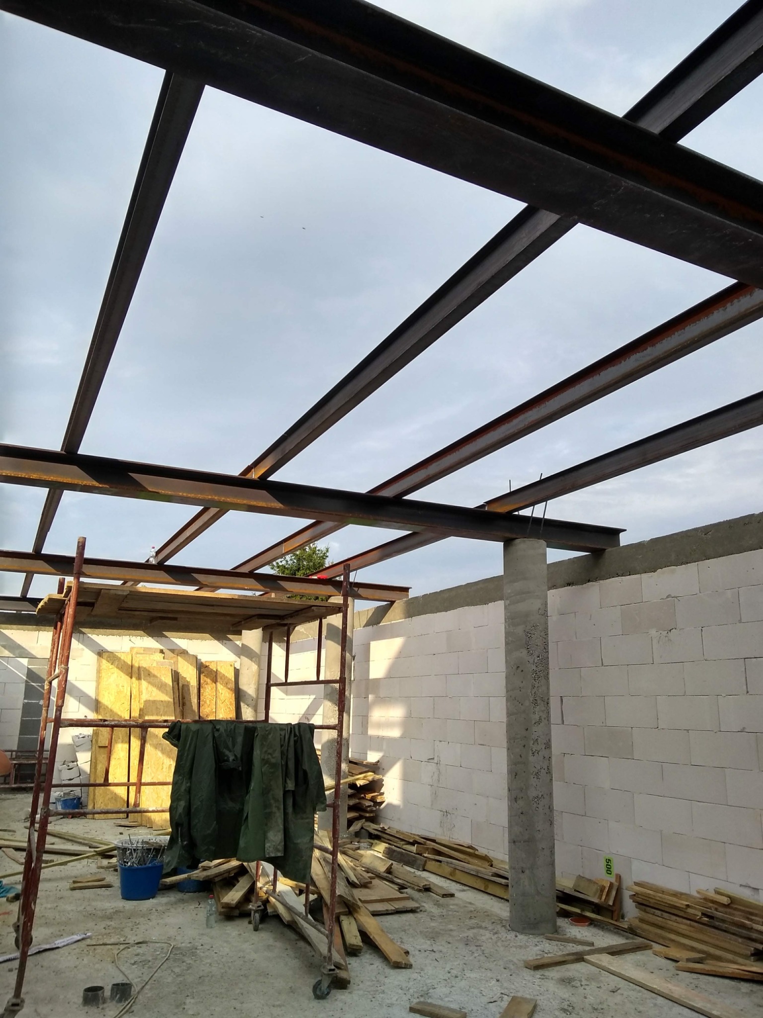 Konstrukcja stalowa dachu w trakcie budowy, widoczne profile stalowe, kolumna betonowa, ściany z betonu komórkowego, rusztowanie i materiały budowlane na placu budowy.