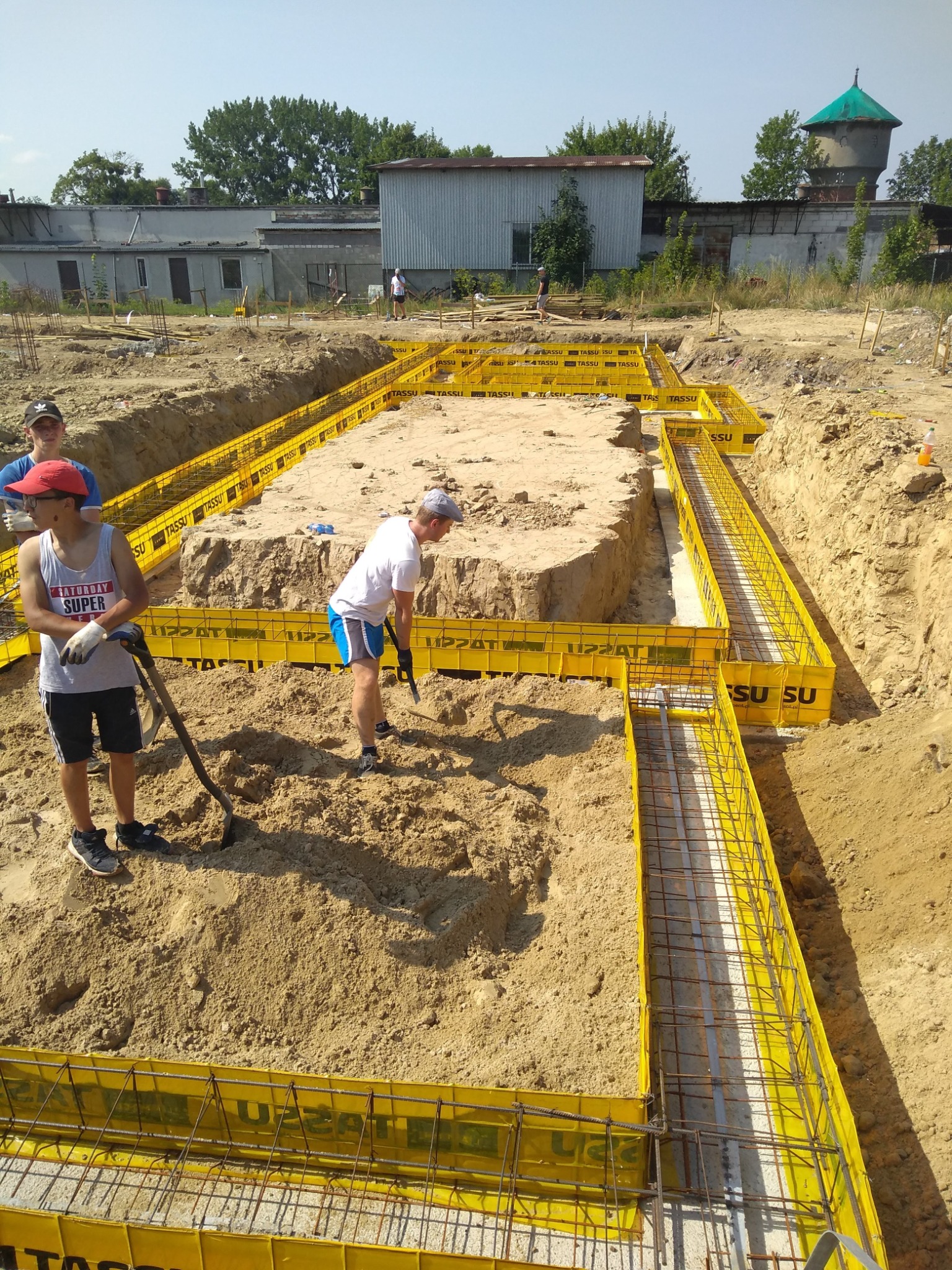 Przygotowanie fundamentów pod budynek: żółte szalunki wypełnione zbrojeniem, pracownicy wyrównują teren łopatami, w tle budynki gospodarcze i wieża ciśnień.