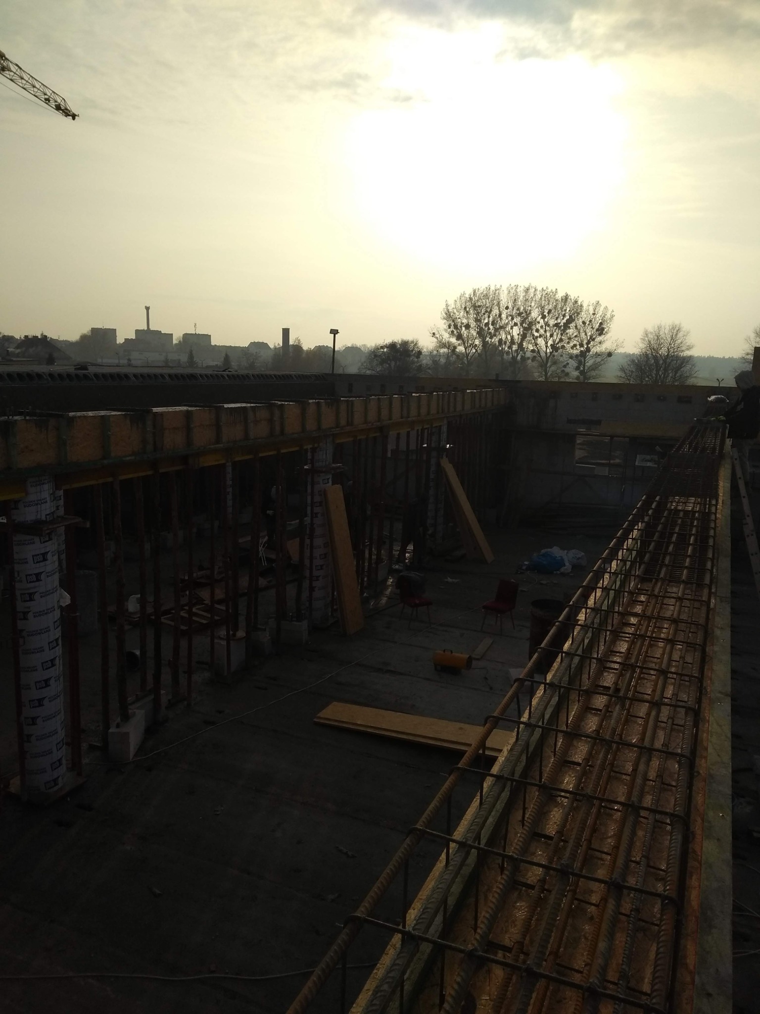 Zbrojenie fundamentów w trakcie budowy, widoczne szalunki, stalowe pręty, narzędzia i krzesła na placu budowy w pochmurny dzień.