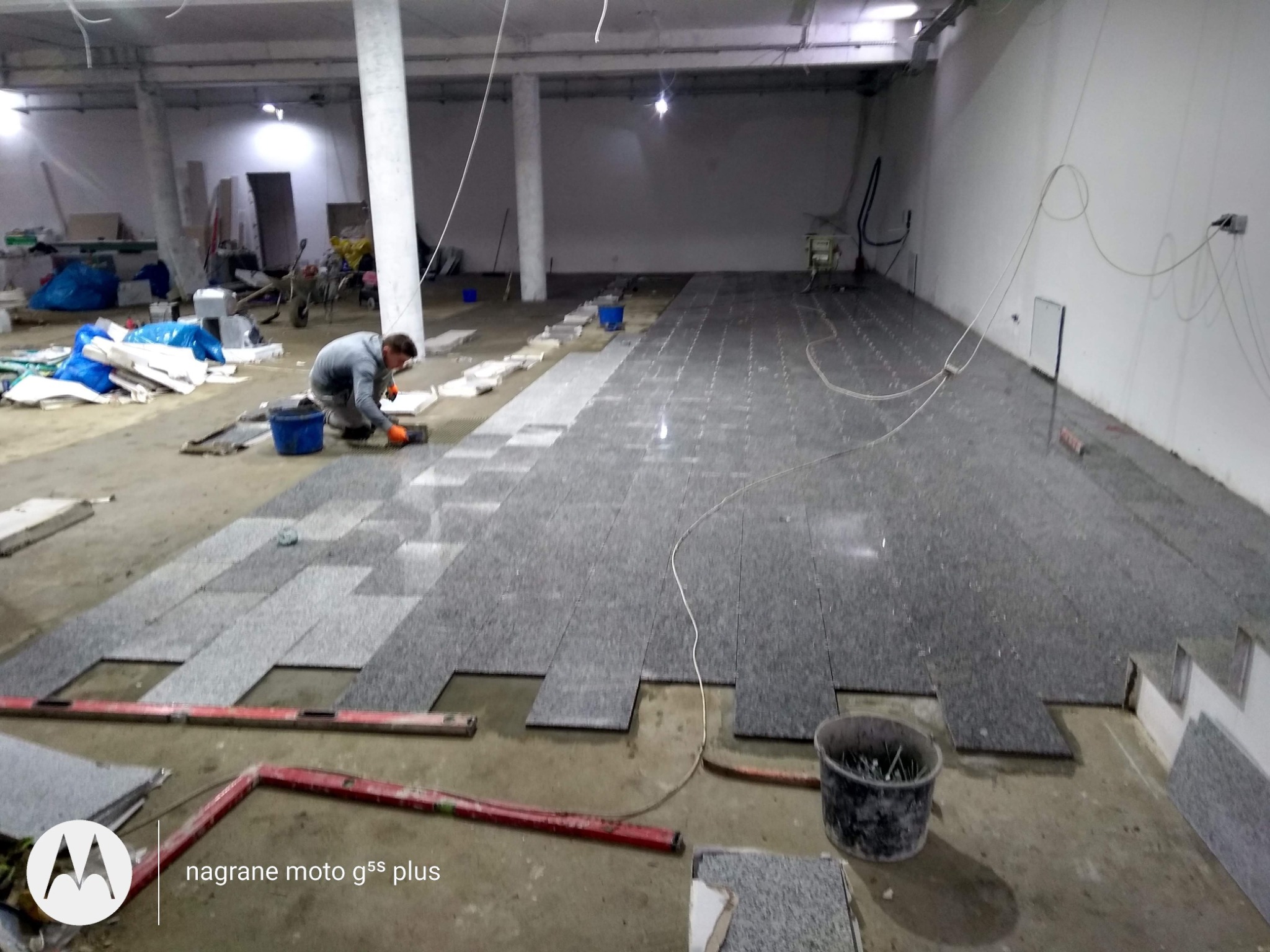 Układanie szarej, granitowej podłogi w dużym, surowym pomieszczeniu; widoczny pracownik w pomarańczowych rękawicach, poziomica i wiadro z narzędziami.