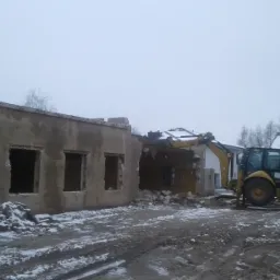 Żółta koparko-ładowarka CAT podczas rozbiórki budynku w pochmurny dzień. Fragmenty gruzu i śniegu na ziemi, pracownik w odblaskowej kamizelce w tle.