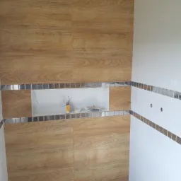 Wykończona ściana łazienki z płytek imitujących drewno, z wnęką dekoracyjną obramowaną mozaiką i białymi płytkami z pasem mozaiki.