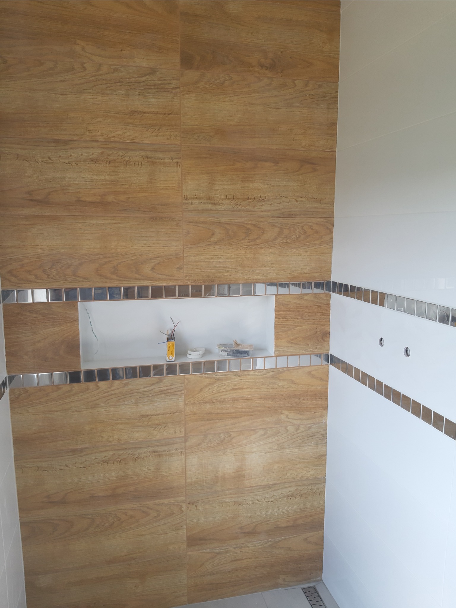 Wykończona ściana łazienki z płytek imitujących drewno, z wnęką dekoracyjną obramowaną mozaiką i białymi płytkami z pasem mozaiki.