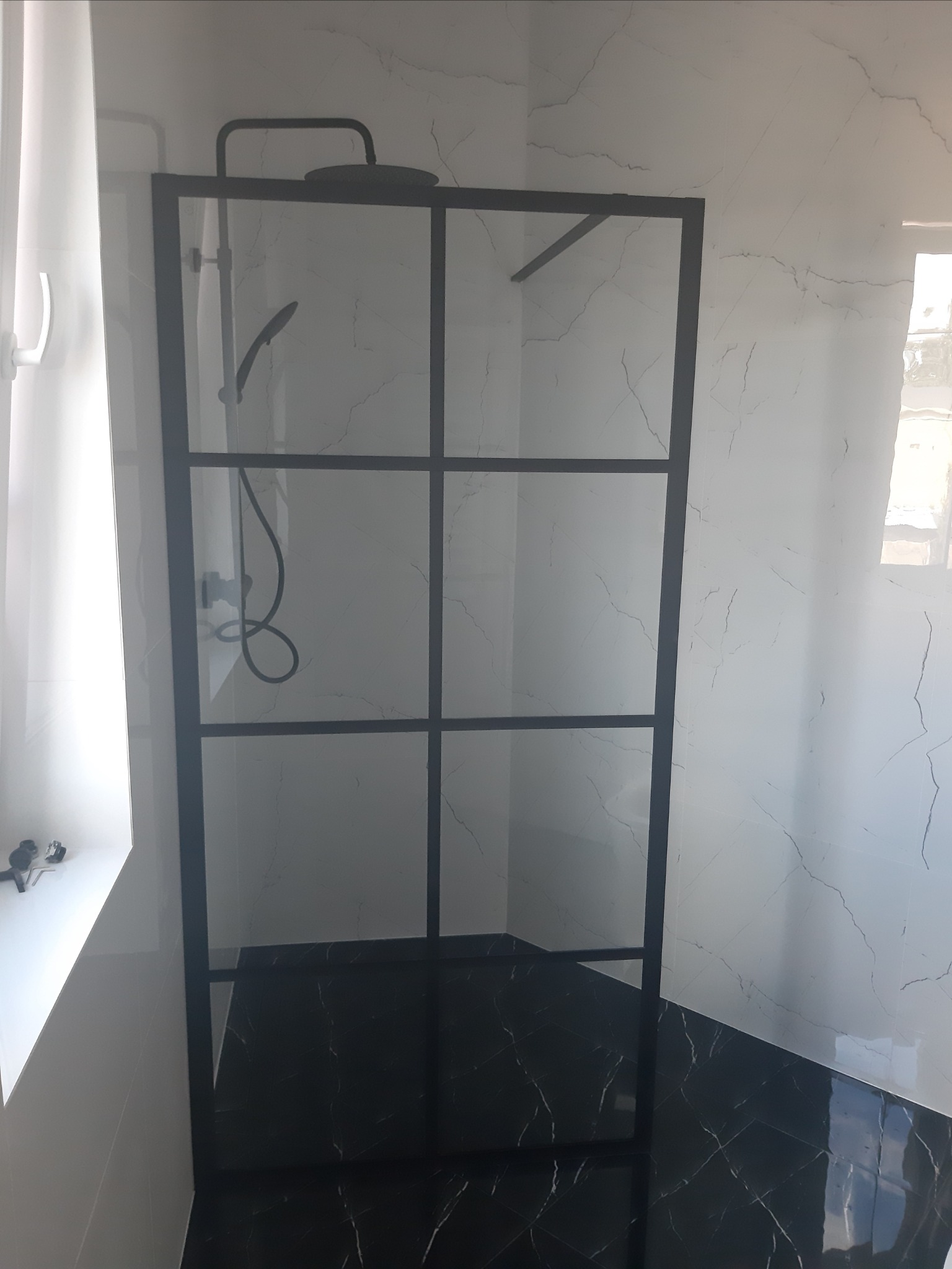 Kabina prysznicowa z czarnymi profilami na tle białych ścian z marmurowym wzorem i czarnej podłogi z motywem marmuru.