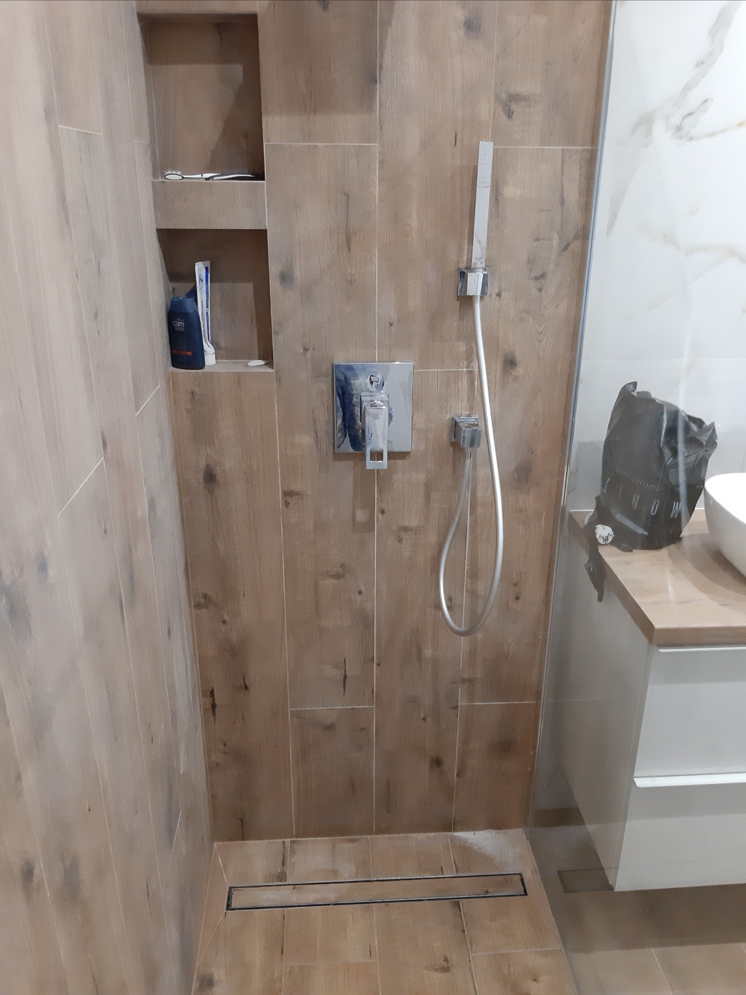 Nowoczesna kabina prysznicowa z drewnopodobnymi płytkami, minimalistyczną armaturą i wnęką z kosmetykami, widoczny odpływ liniowy w podłodze.