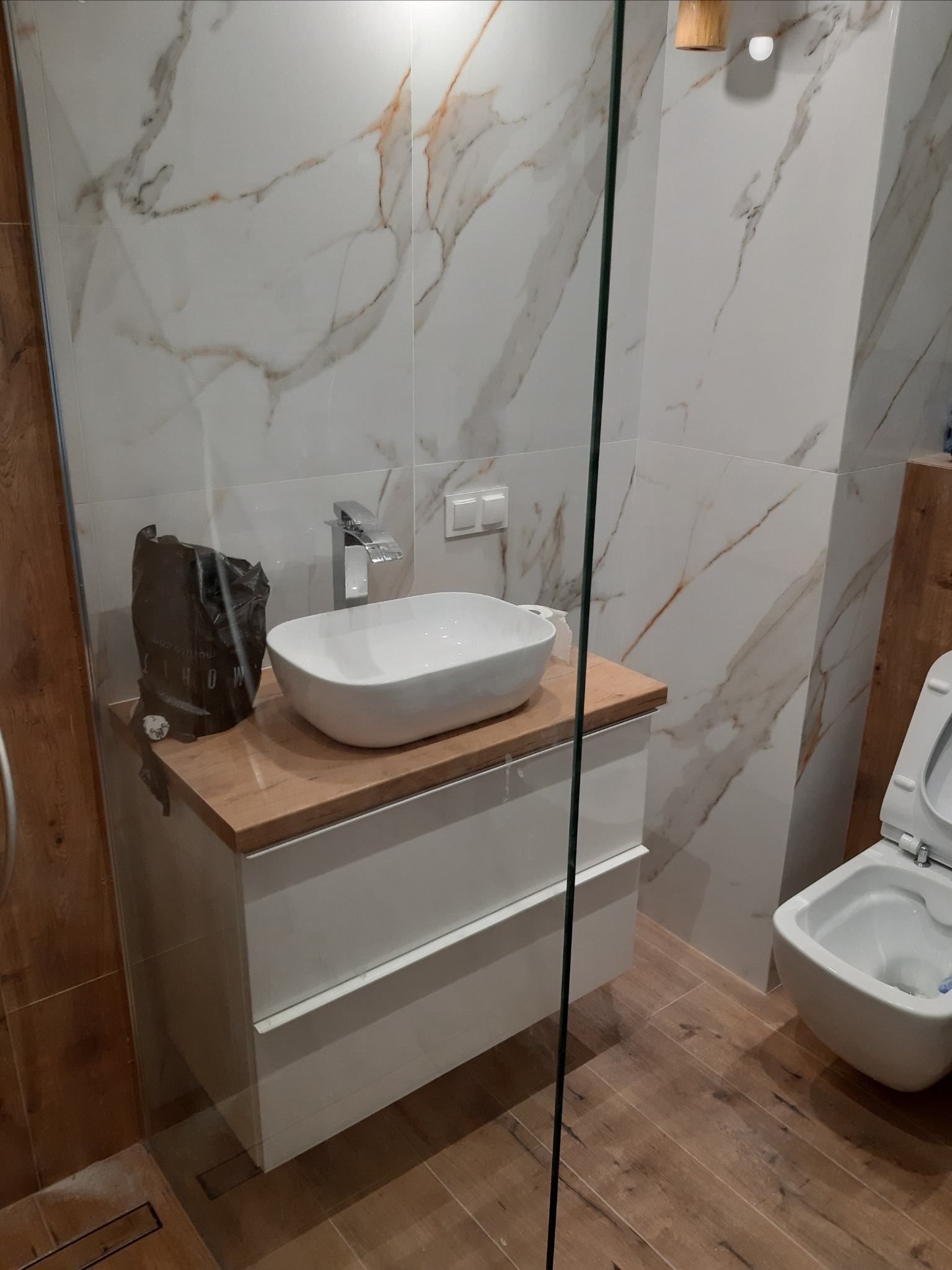 Nowoczesna łazienka z marmurowymi płytkami, umywalką nablatową na szafce z drewnianym blatem i toaletą wiszącą, widok przez szklaną kabinę prysznicową.