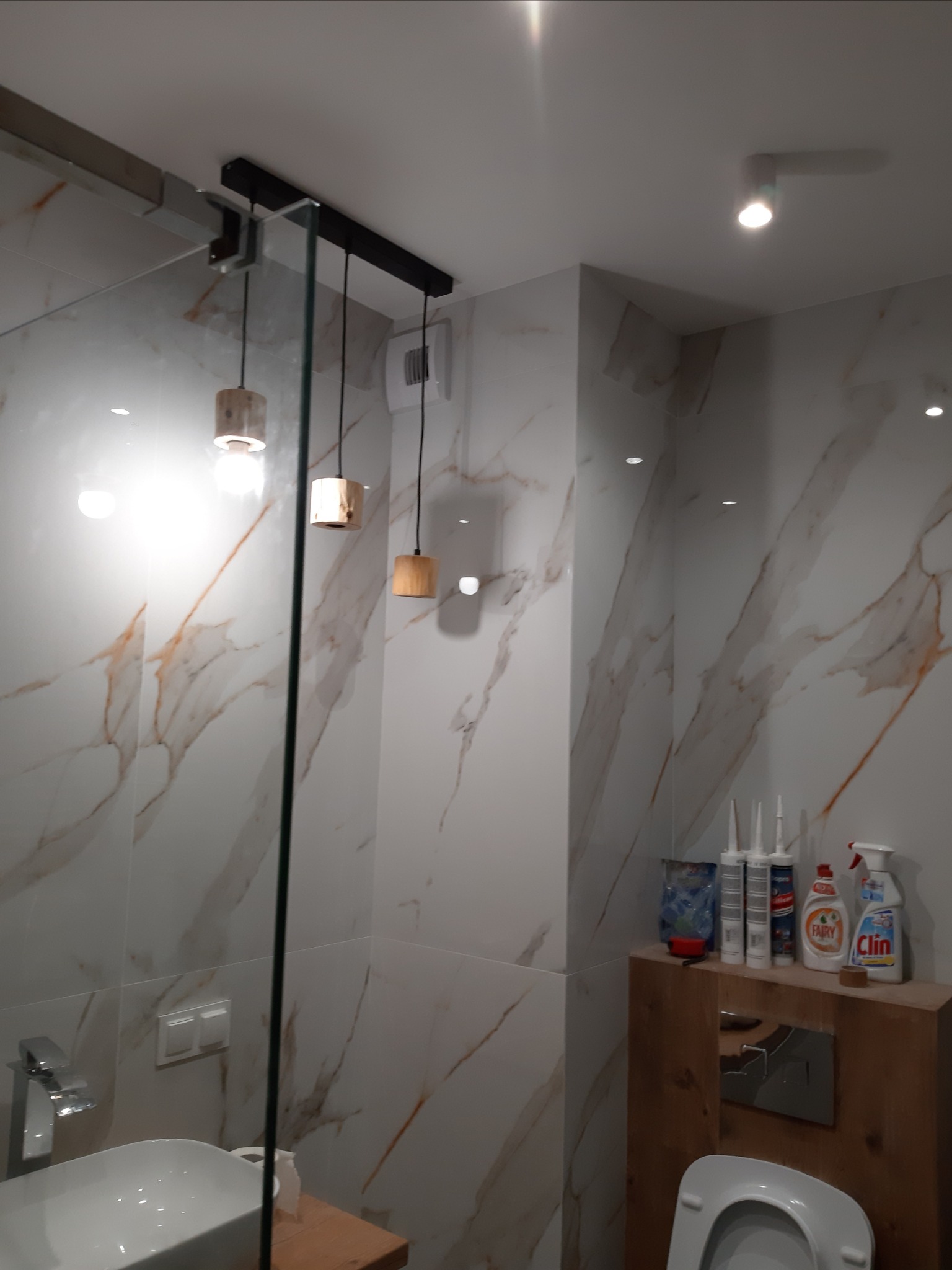 Nowoczesna łazienka z marmurowymi płytkami, wiszącą szafką z drewnianym blatem, umywalką i trzema lampami wiszącymi z drewnianymi elementami. Na szafce widoczne środki czystości i silikony.