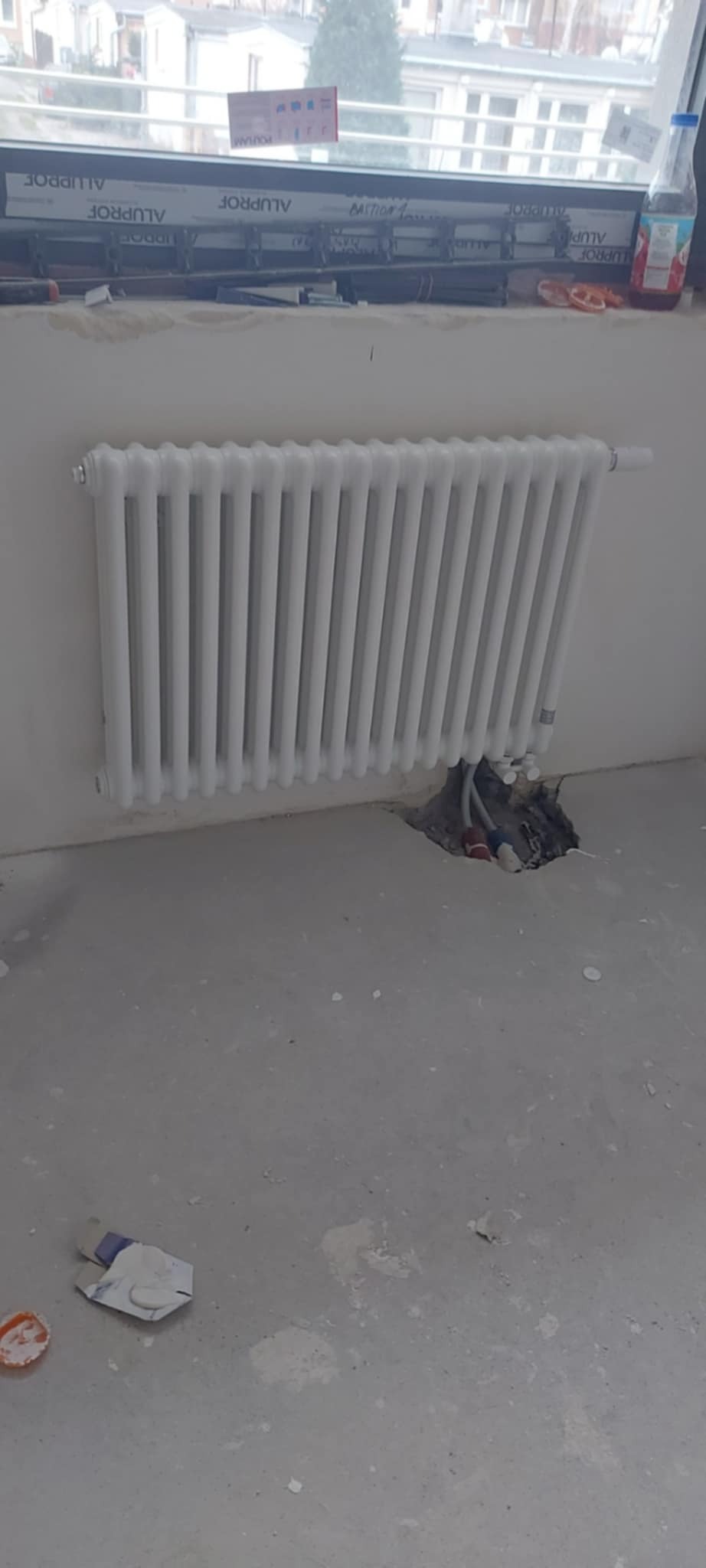 Biały, żeliwny grzejnik zamontowany przy ścianie w trakcie remontu, widoczne przewody w podłodze i niedokończone wykończenie okna.