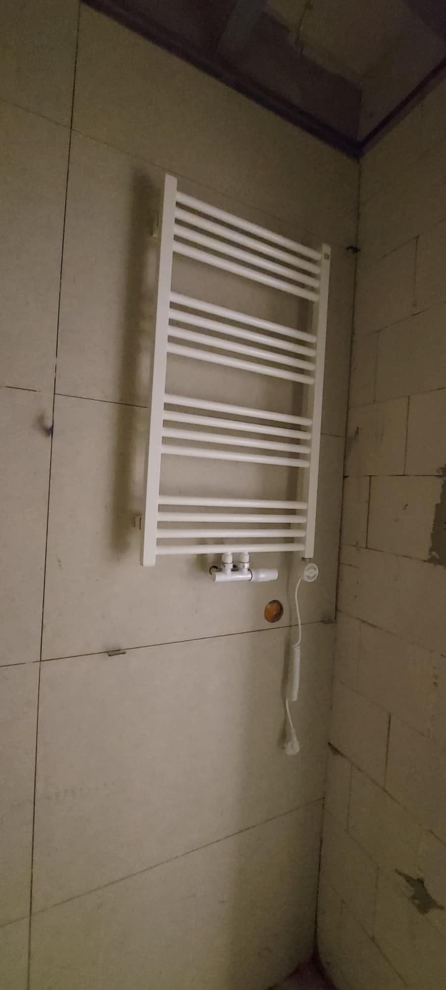 Biały grzejnik łazienkowy drabinkowy zamontowany na ścianie z płytek w trakcie remontu, widoczne przyłącza i kabel termostatu, ściana z niedokończonymi fugami.