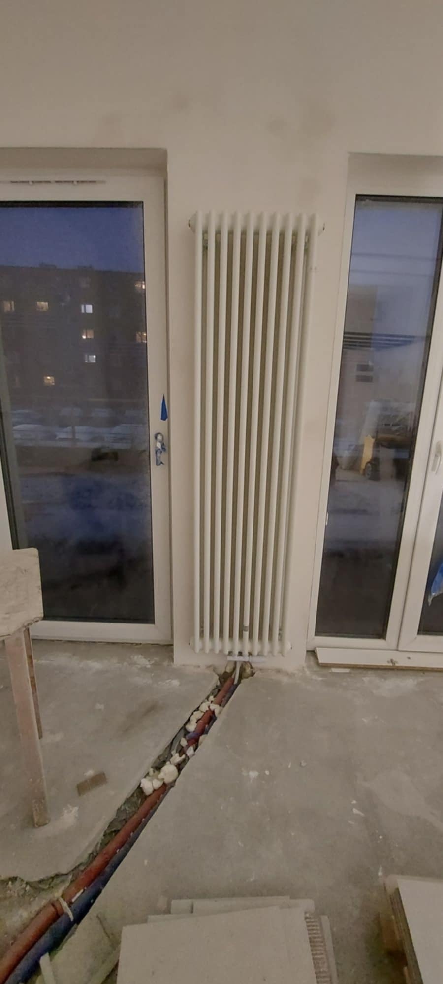 Montaż nowego, białego grzejnika panelowego między oknami balkonowymi, widoczne rury w bruździe w posadzce wypełnione pianką montażową.
