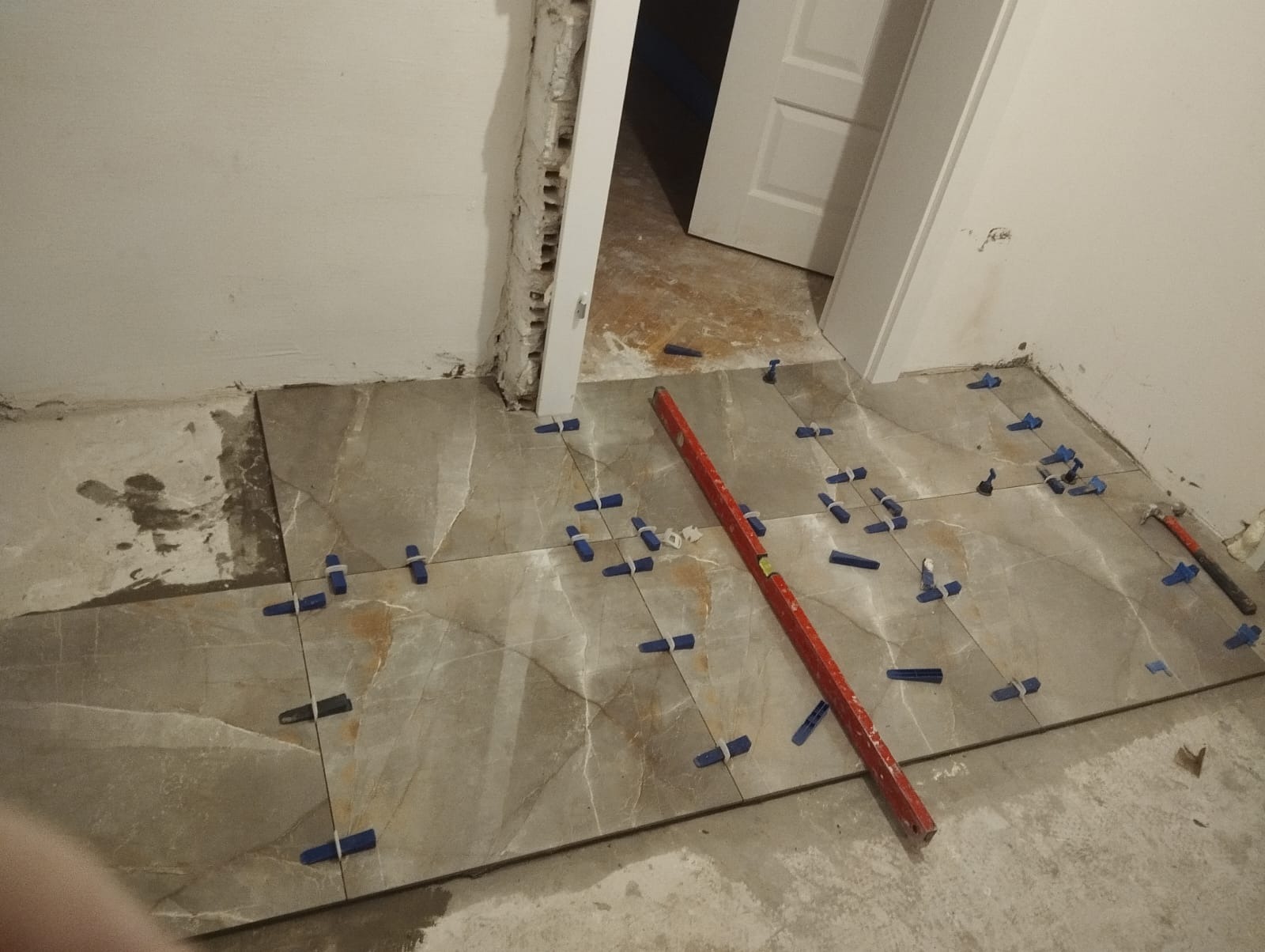 Układanie płytek podłogowych z użyciem poziomicy i klipsów poziomujących w trakcie remontu łazienki, widoczne niedokończone ściany i futryna drzwi.