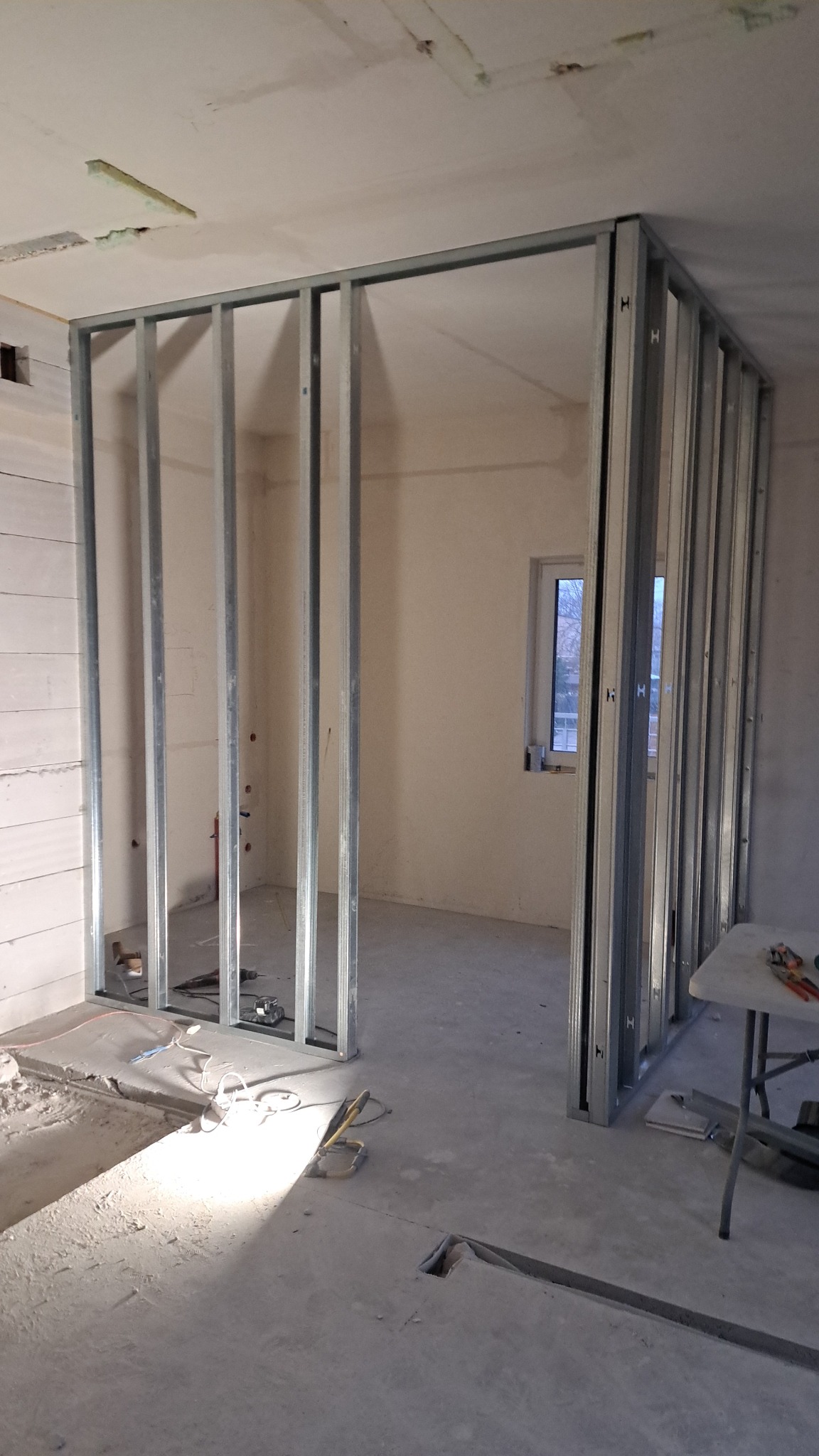 Metalowa konstrukcja szkieletowa ścian działowych w surowym stanie, widoczne przewody, fragment ściany z białych bloczków i okno w tle.