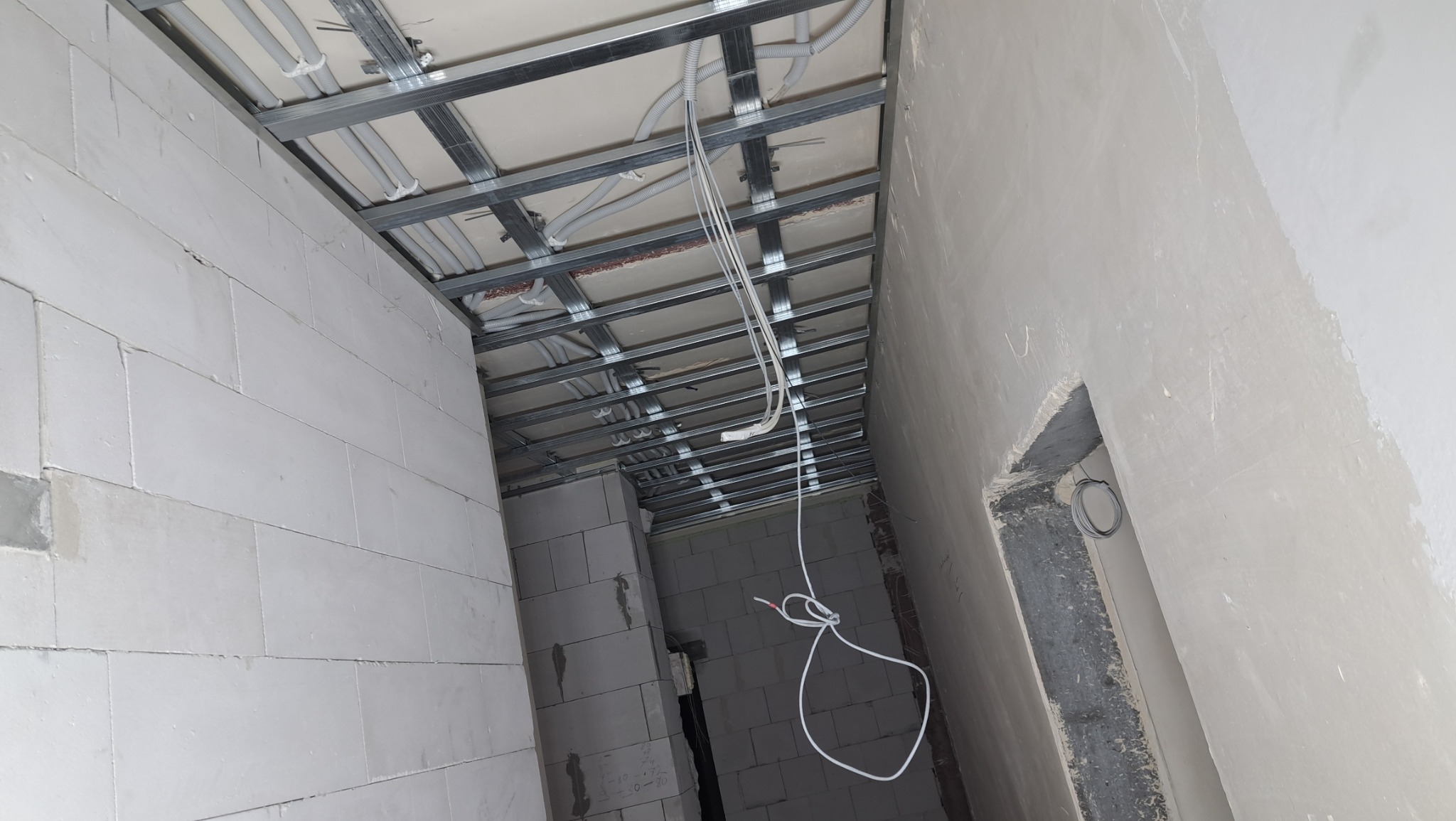 Sufit podwieszany z metalową konstrukcją, widoczne przewody elektryczne i rury wentylacyjne, ściany z bloczków białych i częściowo otynkowane, etap prac wykończeniowych.