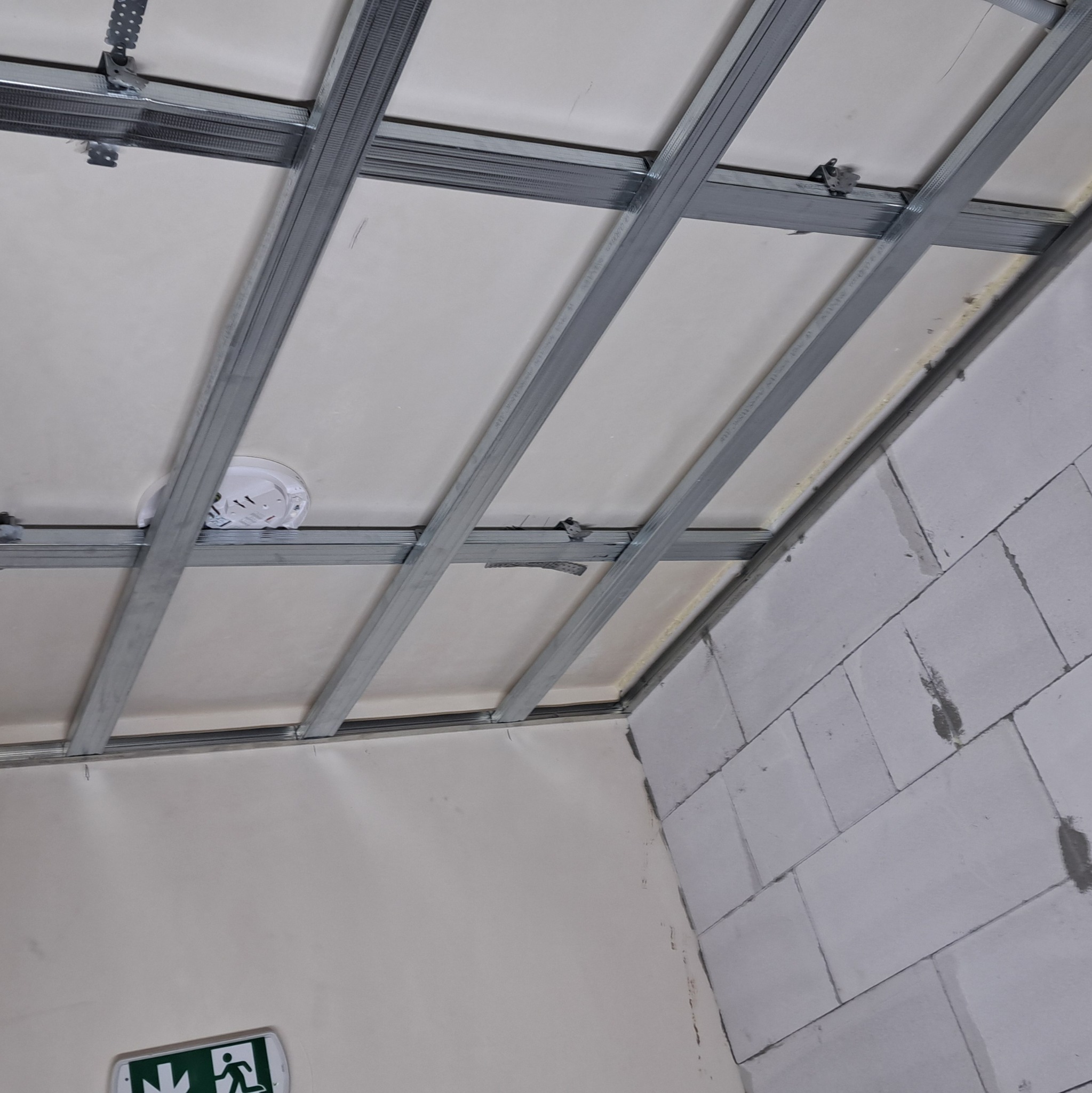 Konstrukcja sufitu podwieszanego z metalowych profili i płyt gipsowo-kartonowych, widoczny czujnik dymu i fragment ściany z białych bloczków.