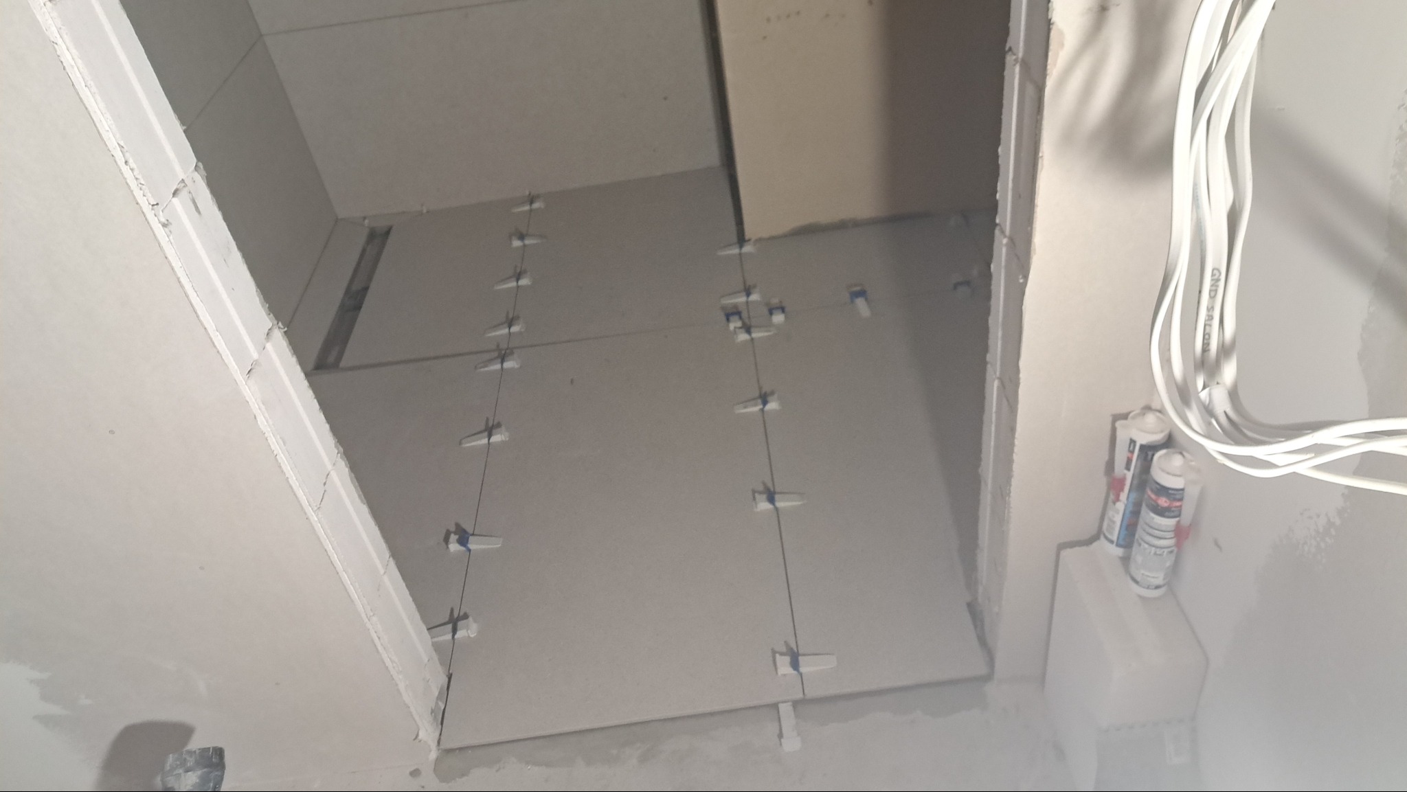 Układanie płytek podłogowych w łazience, widoczne białe krzyżyki dystansowe, przewody elektryczne i tuby z silikonem.