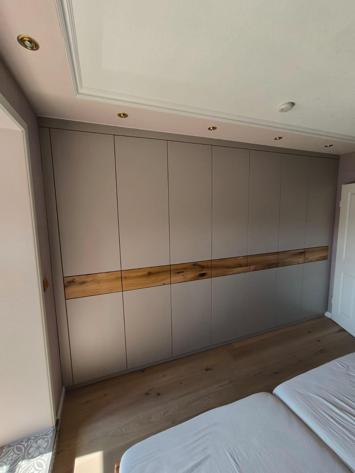 Duża, szara szafa wnękowa z drewnianą wstawką na środku, oświetlenie punktowe na suficie, parkiet na podłodze, widok z dołu na szafę w sypialni.