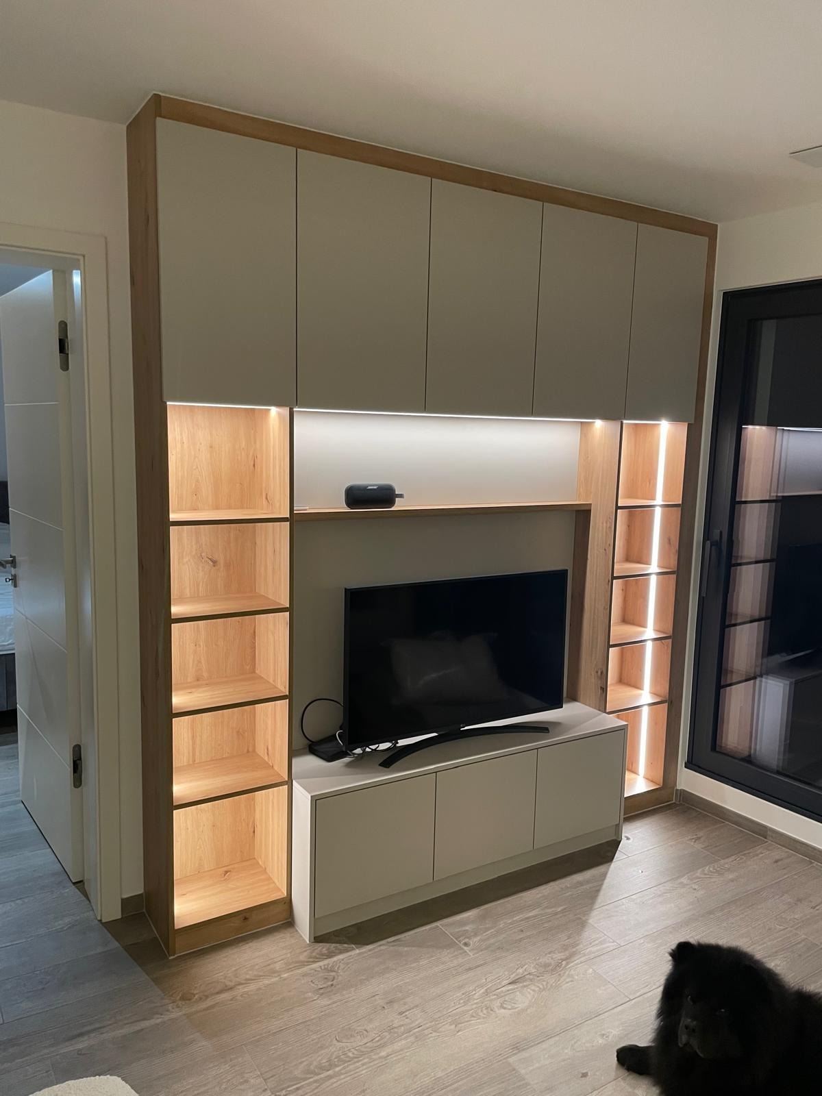 Zabudowa ściany z szafkami wiszącymi i półkami z podświetleniem LED, telewizor na szafce RTV, widoczny fragment podłogi i pies rasy Chow Chow