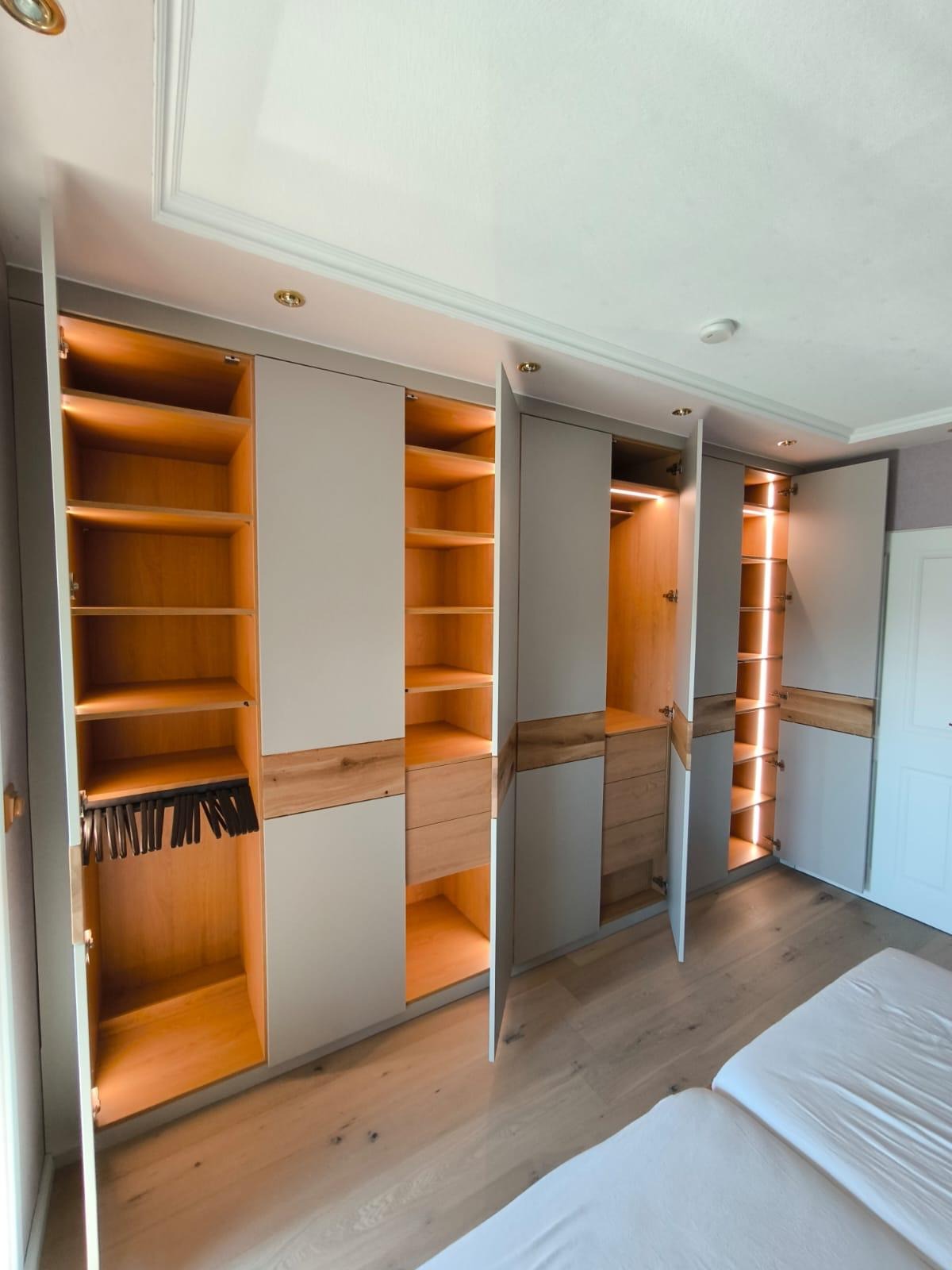 Szara szafa wnękowa z drewnianymi akcentami i oświetleniem LED, częściowo otwarta, z widocznymi półkami i wieszakami na ubrania, na tle jasnej podłogi i łóżka.