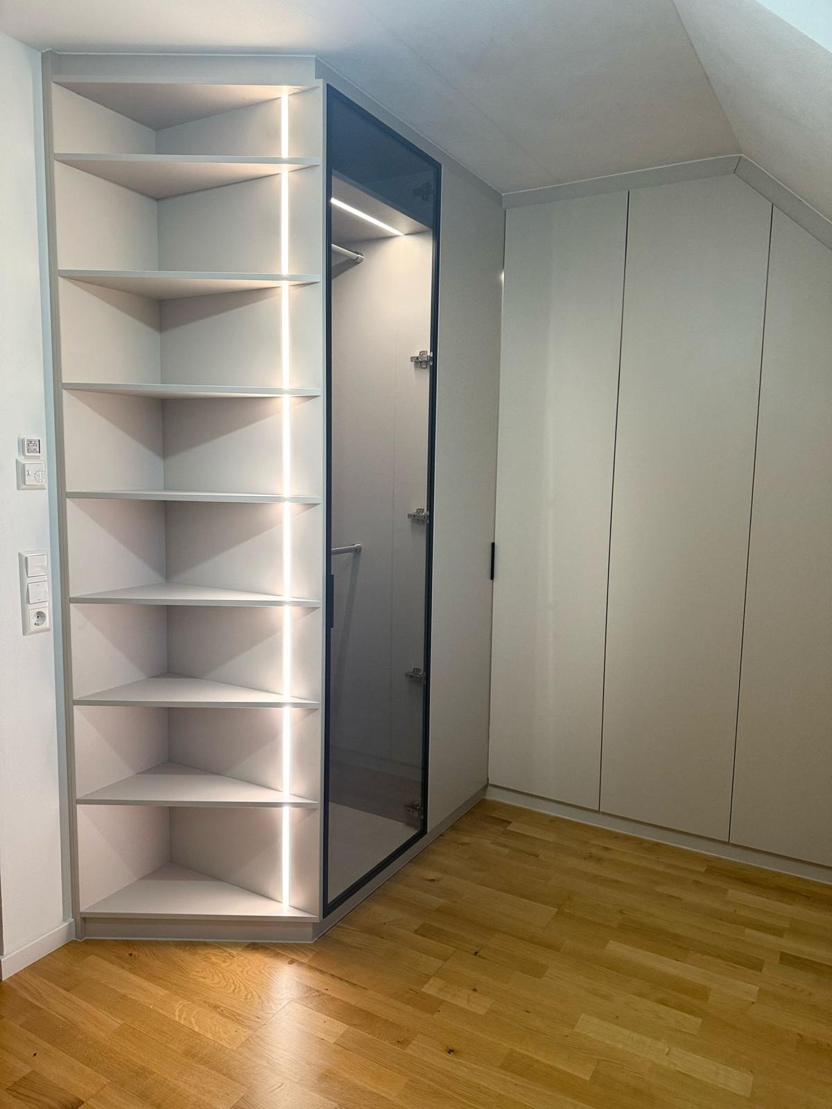 Biała szafa wnękowa z otwartymi półkami narożnymi i podświetleniem LED, drzwi przesuwne z ciemnego szkła, parkiet na podłodze, zabudowa pod skosem dachu.