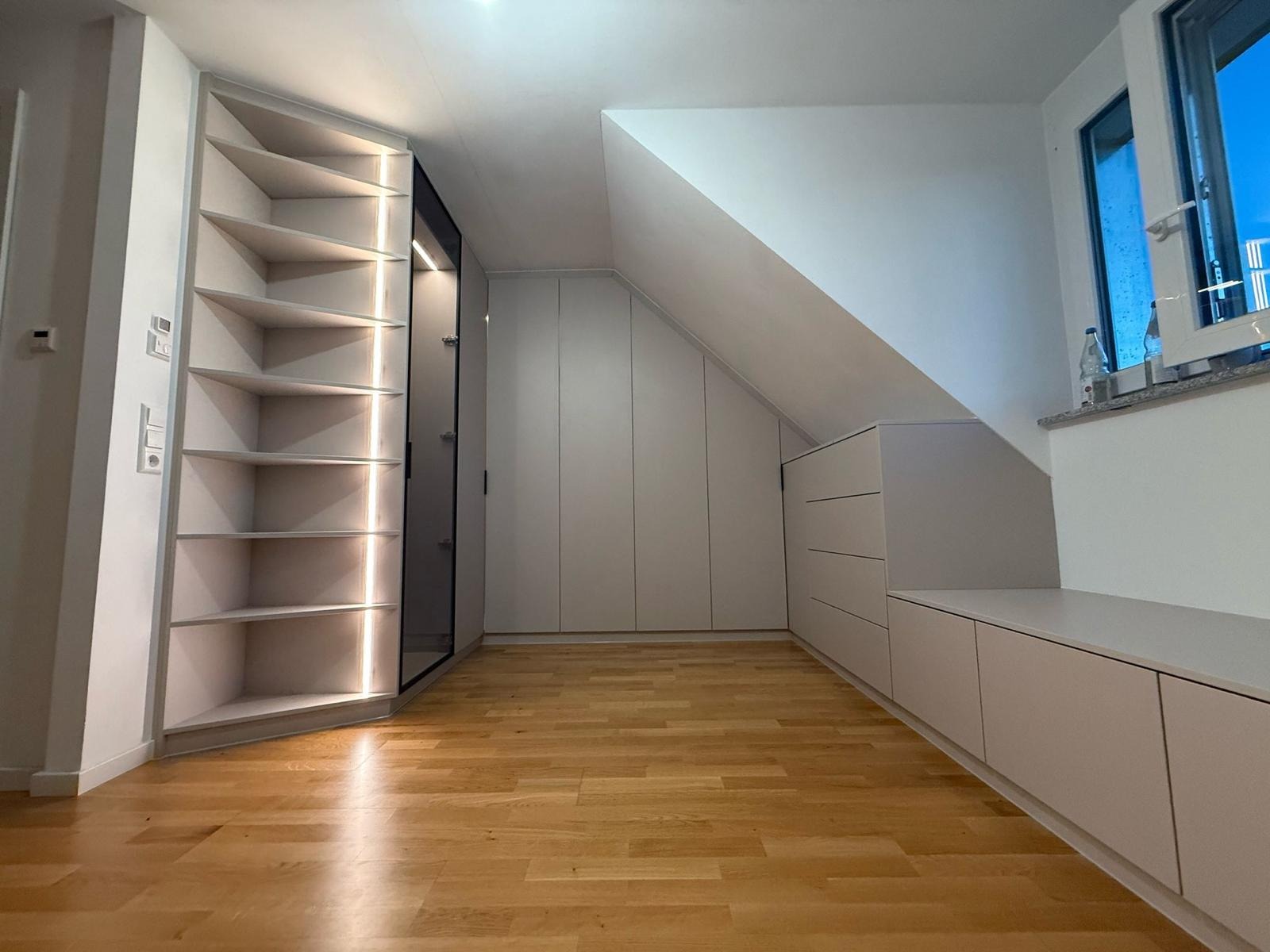 Jasne, dopasowane do skosu poddasza szafy z frontami bez uchwytów i półki z oświetleniem LED, drewniana podłoga.