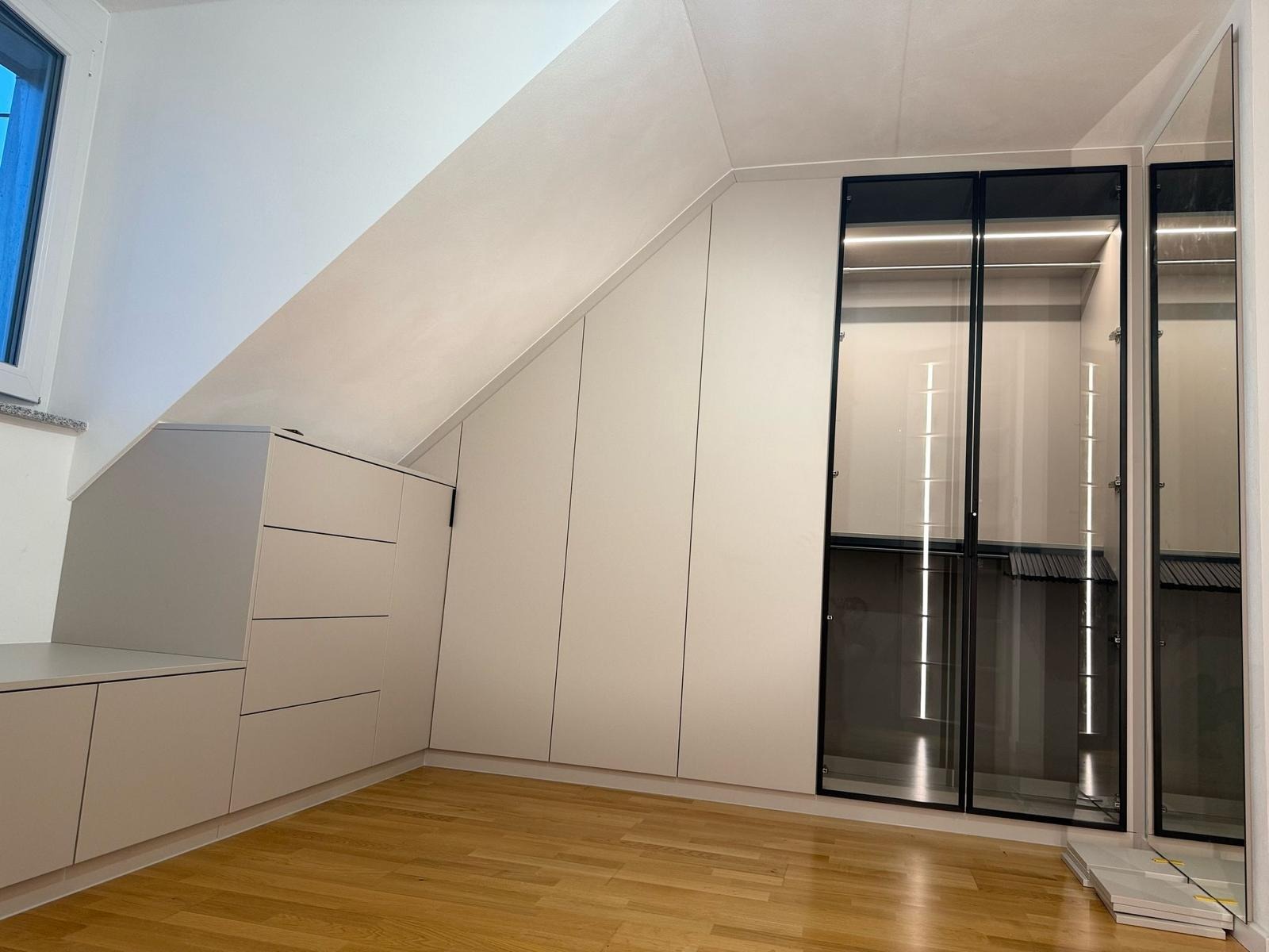 Biała szafa wnękowa z przeszklonymi drzwiami i oświetleniem LED wkomponowana w skos dachu, uzupełniona komodą z szufladami i blatem, na drewnianej podłodze.