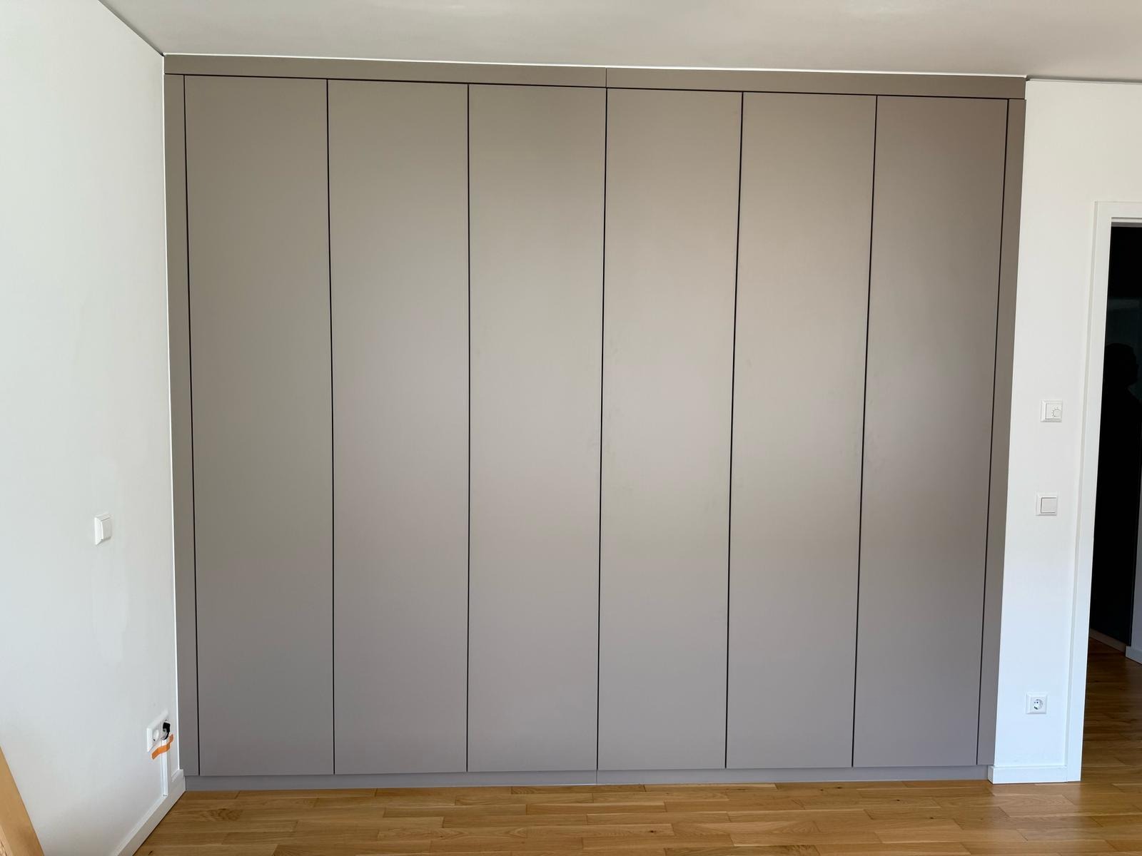 Szafa wnękowa z przesuwnymi drzwiami w odcieniach szarości, zamontowana od podłogi do sufitu, z widocznymi gniazdkami elektrycznymi na ścianie w tle.