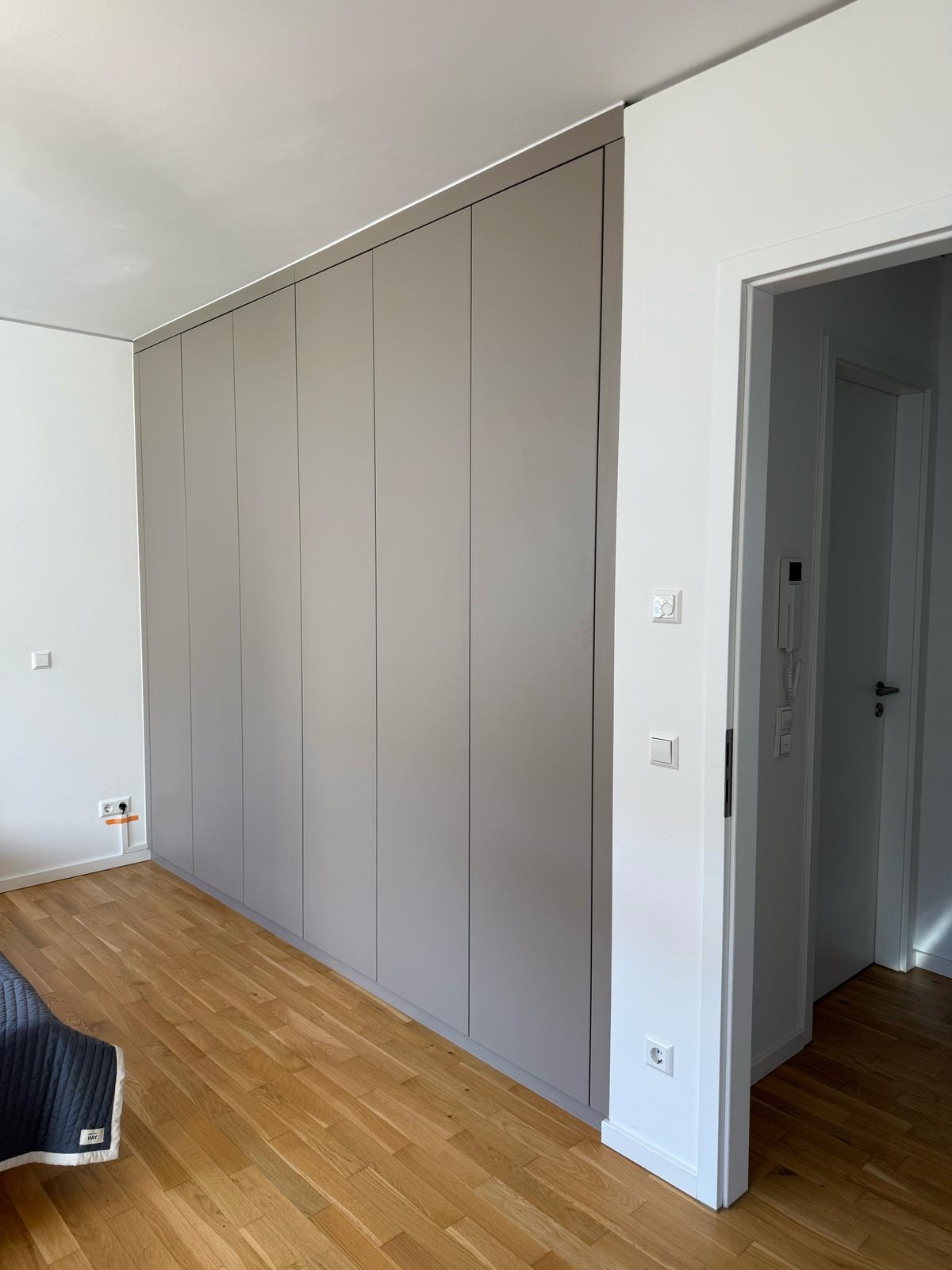 Duża, szara szafa wnękowa z przesuwnymi drzwiami, idealnie dopasowana do ściany w jasnym pokoju z drewnianą podłogą; widoczny fragment łóżka i drzwi wejściowe.