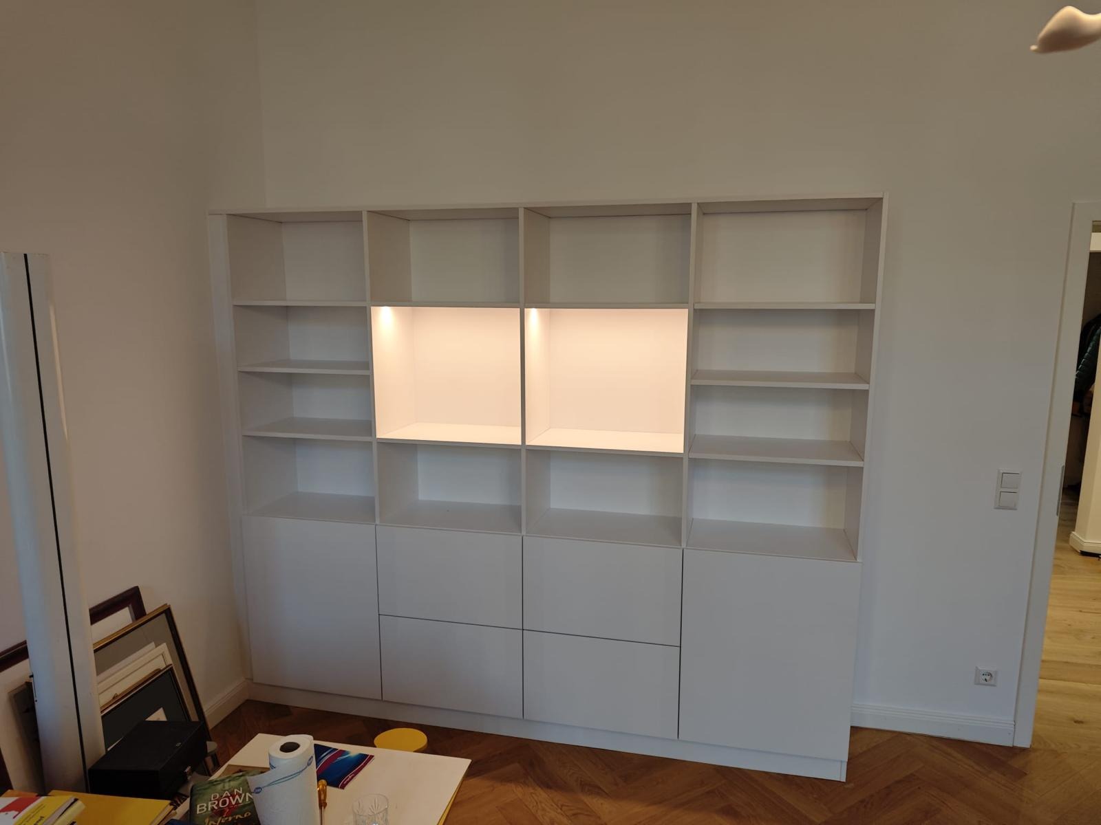 Biała, zabudowana szafa wnękowa z podświetlanymi półkami w centralnej części, na drewnianej podłodze, z widocznymi elementami dekoracyjnymi i ramkami zdjęć w dolnym lewym rogu.