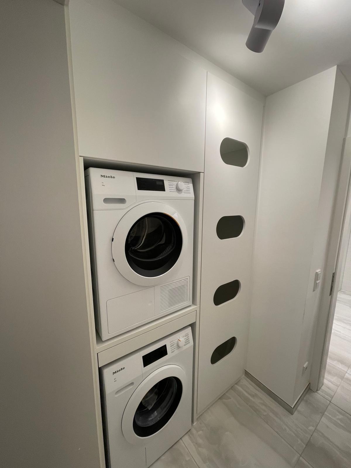 Biała pralka i suszarka Miele w zabudowie meblowej z szafkami z owalnymi otworami wentylacyjnymi, minimalistyczna łazienka z szarymi płytkami podłogowymi.