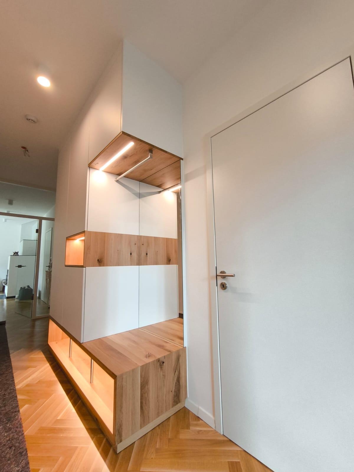 Nowoczesna zabudowa przedpokoju z białymi szafkami i drewnianymi elementami, podświetlana ciepłym światłem LED, widok na parkiet ułożony w jodełkę.