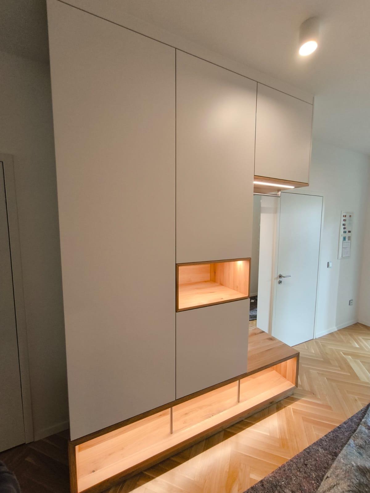 Biała zabudowa meblowa z drewnianymi akcentami i wnękami podświetlonymi ciepłym światłem, widoczna na tle parkietu ułożonego w jodełkę i białych drzwi.