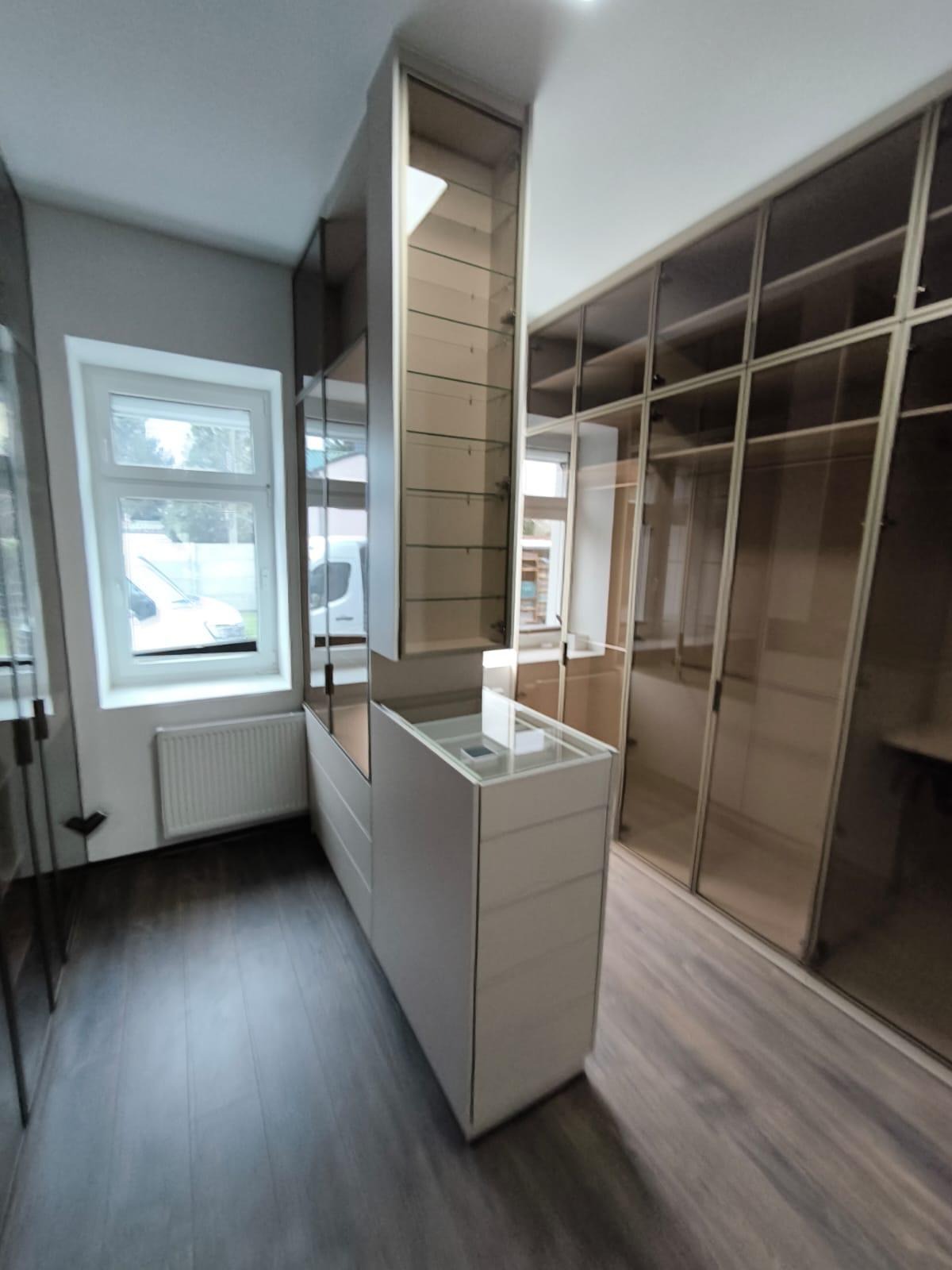 Nowoczesna szafa wnękowa z przesuwnymi drzwiami z brązowego szkła i szklanymi półkami, uzupełniona białą komodą z szufladami, w jasnym pokoju z oknem i grzejnikiem.