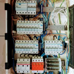 Otwarta skrzynka elektryczna z zamontowanymi bezpiecznikami, ogranicznikami przepięć (kolor pomarańczowy) i licznymi przewodami w kolorach niebieskim, brązowym, żółto-zielonym, zamontowana...