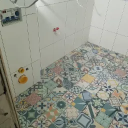 Wnętrze łazienki w trakcie remontu: kolorowe płytki podłogowe z wzorami, białe płytki ścienne, instalacja podtynkowa i wystające przewody elektryczne.