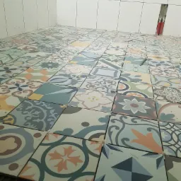 Układanie patchworkowej podłogi z płytek ceramicznych w łazience, widoczne krzyżyki dystansowe i fragment ściany z instalacją hydrauliczną.