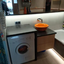 Nowoczesna łazienka z pralką pod blatem, pomarańczowa umywalka w kształcie misy, podświetlenie LED pod szafką i lustro z odbiciem osoby robiącej zdjęcie.