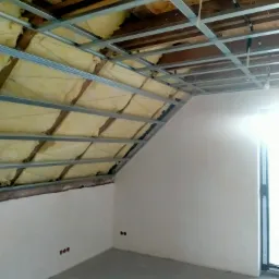 Poddasze z izolacją, metalowa konstrukcja sufitu i gładkie ściany. Widoczne przygotowane punkty elektryczne. Okno wpuszcza jasne światło.