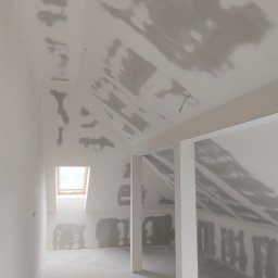 usługi remontowo wykończeniowe Paweł Paprocki - Poddasze ze skosami w trakcie prac wykończeniowych. Widoczne ślady po gładzi na ścianach i suficie. Małe okno dachowe wpuszcza światło.