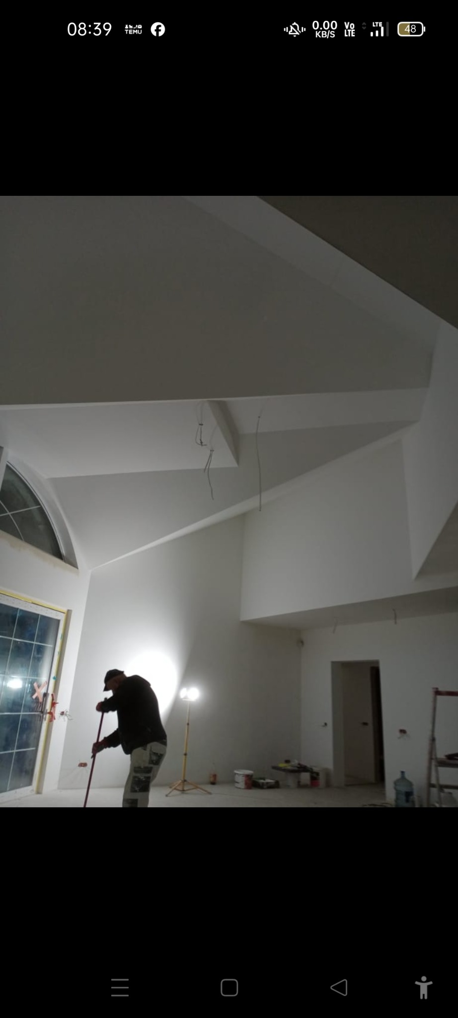 Wysokie wnętrze po gładziowaniu: białe ściany, okno z łukiem, widoczne przewody elektryczne. Mężczyzna szlifuje podłogę. Scena z remontu.