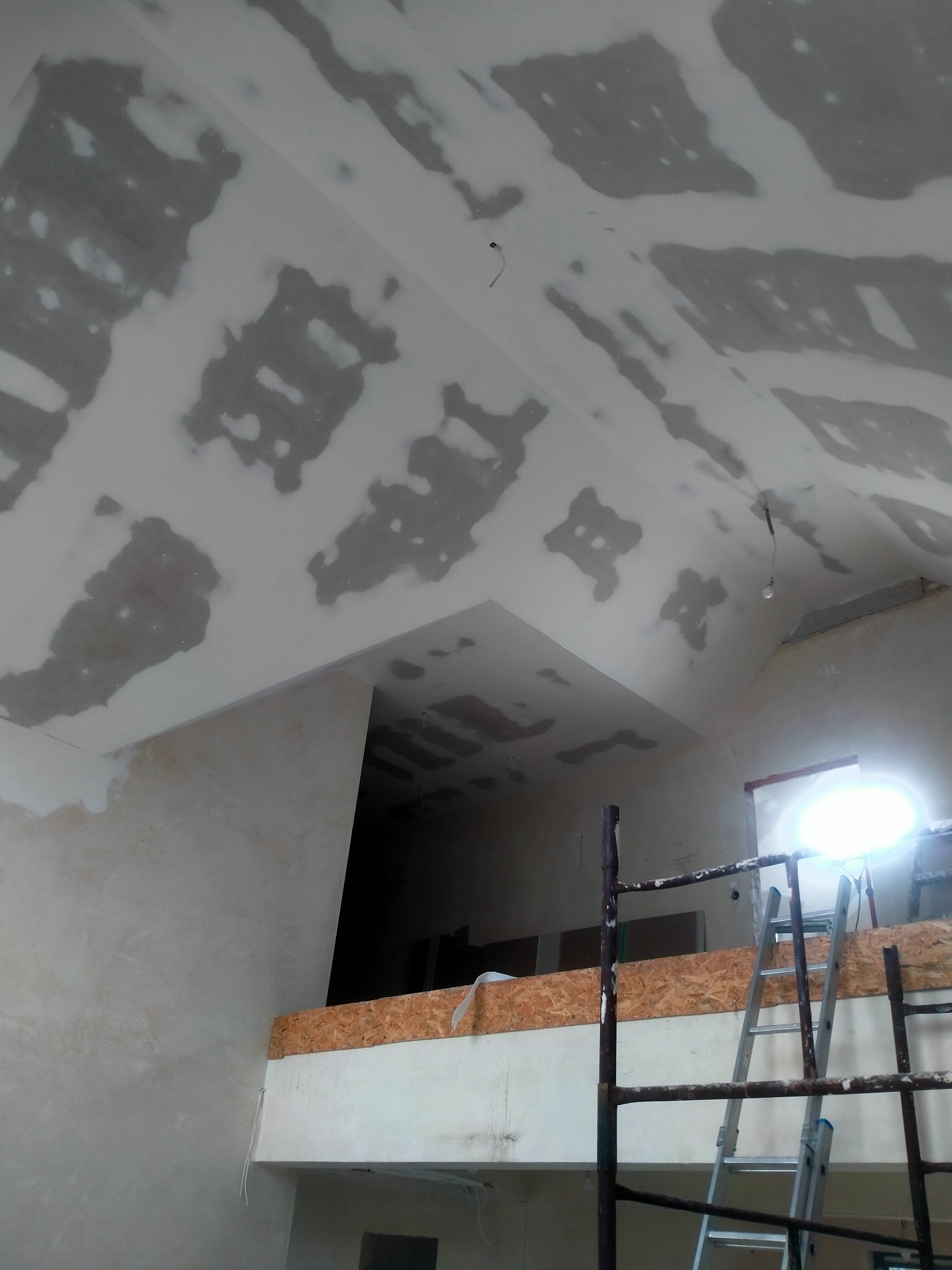Skośny sufit po nałożeniu gładzi szpachlowej, widoczne ślady po łatach. Metalowe rusztowanie i drabina oparta o ścianę. W tle otwór w ścianie.