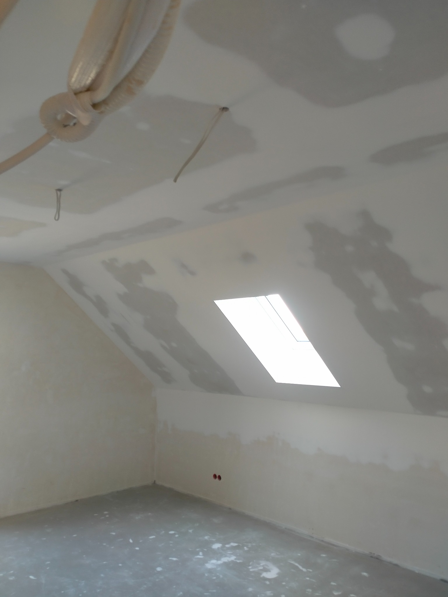 Poddasze ze skośnym oknem dachowym w trakcie prac wykończeniowych; widoczne ślady gładzi na ścianach i suficie, oraz elementy instalacji wentylacyjnej.