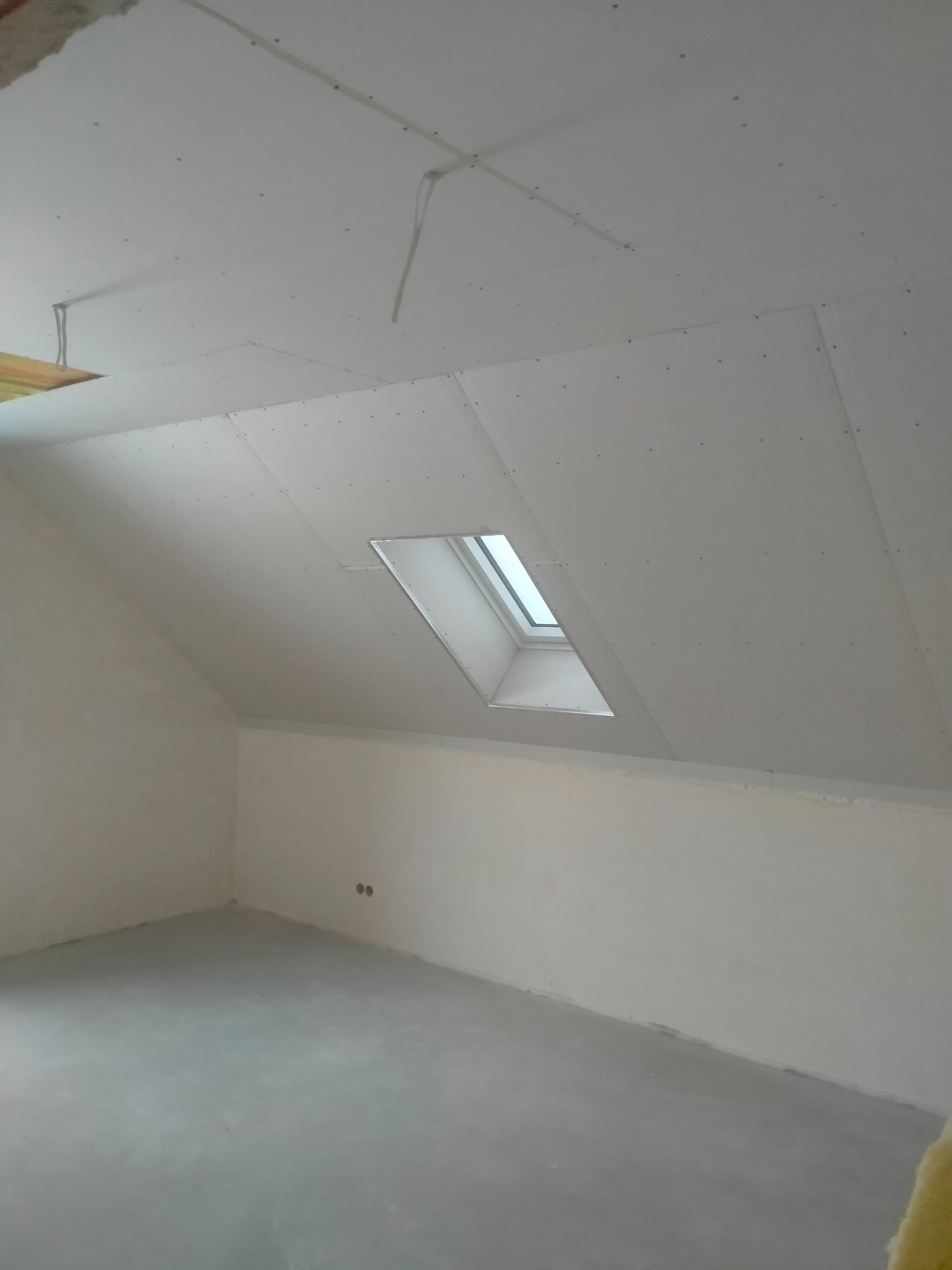 Poddasze ze skośnym sufitem, okno dachowe, płyty karton-gips na suficie, widoczne kable instalacji elektrycznej. Stan surowy, przygotowanie do dalszych prac wykończeniowych.