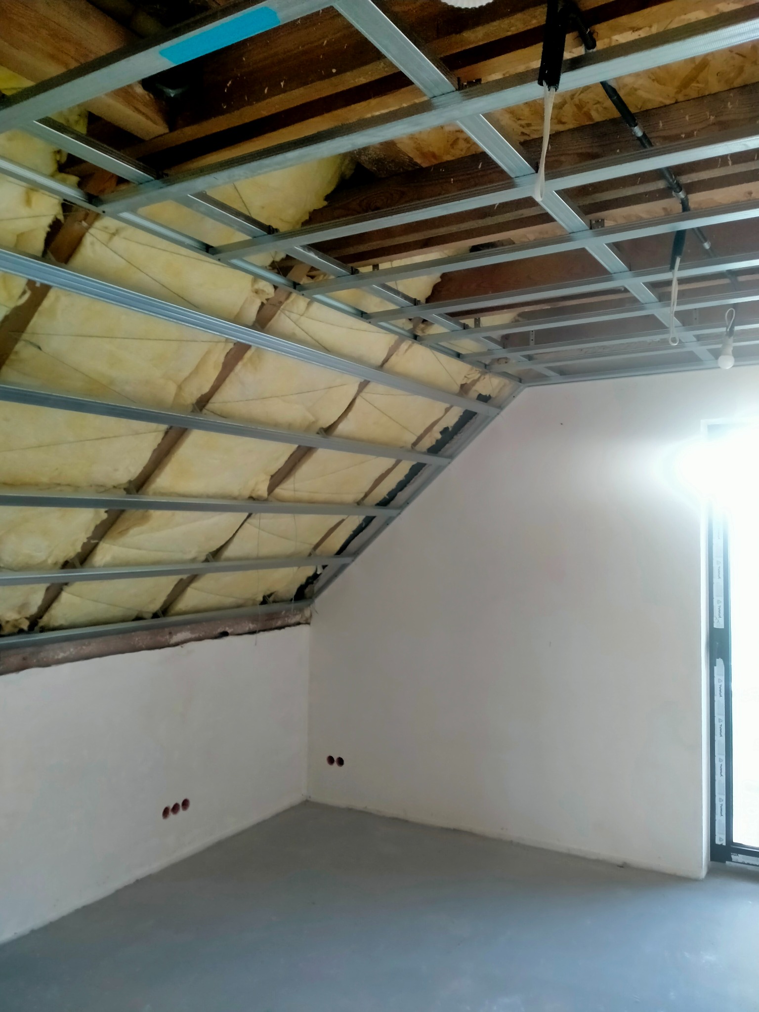 Poddasze z izolacją, metalowa konstrukcja sufitu i gładkie ściany. Widoczne przygotowane punkty elektryczne. Okno wpuszcza jasne światło.