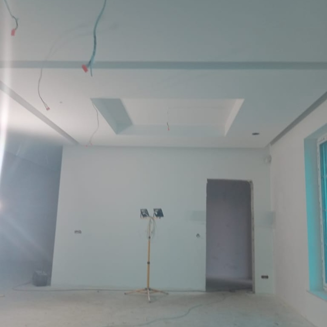 Nowoczesne wykończenie wnętrza: białe ściany z gładzią, sufit podwieszany z oświetleniem. Widoczne kable i halogen na statywie. Okno w tle.