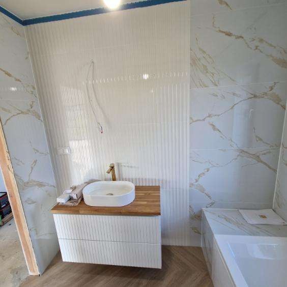 Nowoczesna łazienka z umywalką nablatową na drewnianym blacie, białe płytki imitujące marmur na ścianach i podłodze w jodełkę. Złota bateria i minimalistyczny design.