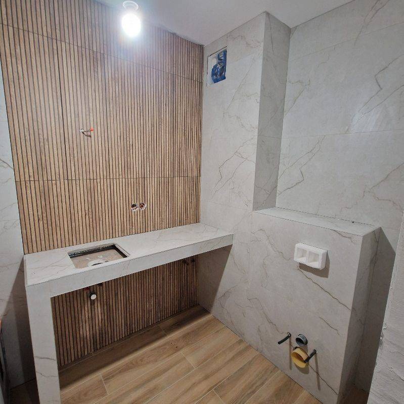 Wykończona łazienka z drewnianą ścianą z lameli, marmurową umywalką i płytkami imitującymi marmur na ścianach. Widoczne instalacje wodne i elektryczne.