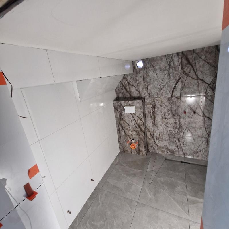 Wykończona łazienka z białymi i marmurkowymi płytkami ściennymi oraz szarą podłogą. Widoczne detale montażowe i odpływ liniowy.