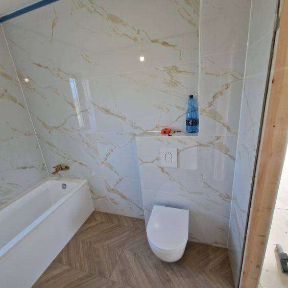 Wykończona łazienka z wanną, toaletą i marmurowymi płytkami w odcieniach bieli i złota. Na półce butelka wody i miarka. Podłoga wyłożona panelami w jodełkę.